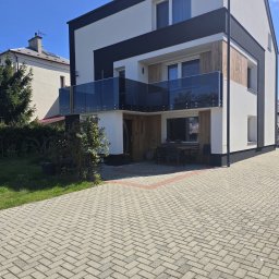mfast - Nowoczesny dom z biało-grafitową elewacją, drewnianymi panelami i przeszkloną barierką balkonu. Przed budynkiem jasna kostka brukowa w jodełkę z czerwoną obwódką.