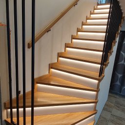 mfast - Nowoczesne drewniane schody zabiegowe z podświetleniem LED pod każdym stopniem, czarnymi pionowymi elementami balustrady i drewnianą poręczą.