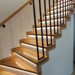 mfast - Nowoczesne, kręcone schody drewniane z dębowymi stopniami, podświetlane listwami LED, z czarnymi, prostokątnymi słupkami balustrady i drewnianą poręczą ścienną.
