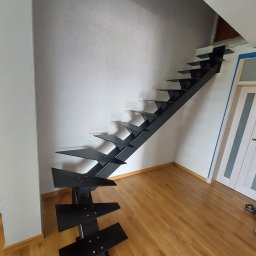 mfast - Czarne, metalowe schody modułowe o nowoczesnej, otwartej konstrukcji z płaskimi stopniami do wykończenia, zamontowane w jasnym wnętrzu z drewnianą podłogą.