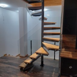 mfast - Eleganckie schody spiralne z trójkątnymi drewnianymi stopniami, ciemnym metalowym słupem i przezroczystą balustradą szklaną, zamontowane w nowoczesnym wnętrzu.