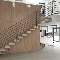 mfast - Nowoczesne schody z jasnymi, drewnianymi stopniami i ciemną, poziomą balustradą metalową, na tle panelowej ściany. W tle widoczna kuchnia i mężczyzna na piętrze.