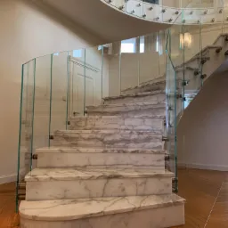 Eleganckie, spiralne schody z marmuru Carrara z przezroczystą balustradą szklaną mocowaną punktowo, widok z dołu na jasne wnętrze.