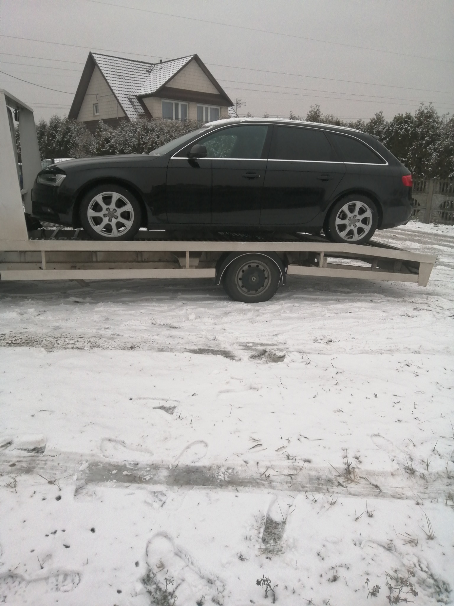 Czarny samochód kombi marki Audi na platformie transportowej w zimowej scenerii, z widocznym domem w tle.