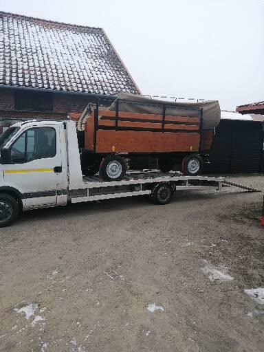 Biały samochód dostawczy z platformą transportową przewożący drewnianą przyczepę z plandeką na tle budynku z dachówką pokrytą śniegiem.