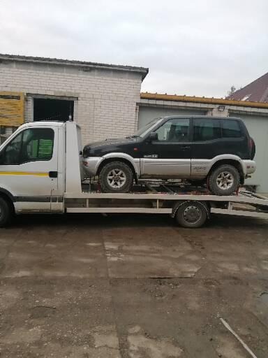 Czarno-srebrny SUV transportowany na białej platformie pomocy drogowej przed budynkiem z cegły w pochmurny dzień.