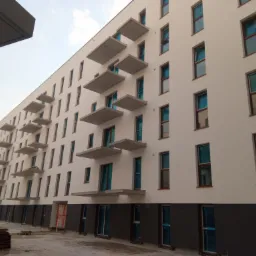 Nowoczesny budynek wielorodzinny z białymi ścianami, szarymi balkonami i oknami zabezpieczonymi folią, widok z dołu w pochmurny dzień.