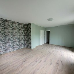 Chamer Maja Hałub - Jasne panele podłogowe w pustym pokoju z zielonymi ścianami i akcentem w postaci ściany z tapetą w kwiaty. Widoczne drzwi i okno.