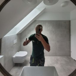 Chamer Maja Hałub - Wykończona łazienka na poddaszu w szarościach, widok w lustrze. Ukończona toaleta podwieszana, płytki na ścianach i podłodze. Fachowiec robi selfie, kciuk w górę.