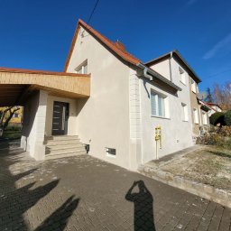 Chamer Maja Hałub - Odnowiona elewacja domu jednorodzinnego z jasnym tynkiem, widoczne detale architektoniczne i podjazd z kostki brukowej. Słoneczny dzień, błękitne niebo.