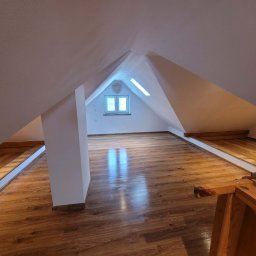 Chamer Maja Hałub - Wykończone poddasze z drewnianą podłogą i białymi ścianami. Widoczne okno dachowe i konstrukcja dachu. Podłoga z wyraźnym wzorem drewna, odbijająca światło.