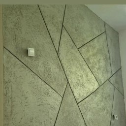 Chamer Maja Hałub - Dekoracyjna ściana z efektem betonu i geometrycznym podziałem na panele. Widoczny włącznik światła i gniazdko elektryczne. Szara kolorystyka.