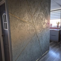 Chamer Maja Hałub - Dekoracyjna ściana w odcieniach szarości z geometrycznym wzorem podkreślonym złotymi liniami, widoczna obok szarych drzwi i fragmentu podłogi.