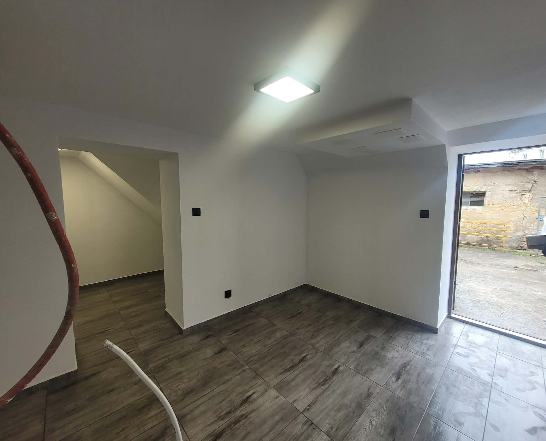 Wykończone wnętrze z panelami podłogowymi imitującymi drewno, świeżo pomalowanymi ścianami i nowoczesnym oświetleniem sufitowym. Widoczne fragmenty instalacji.