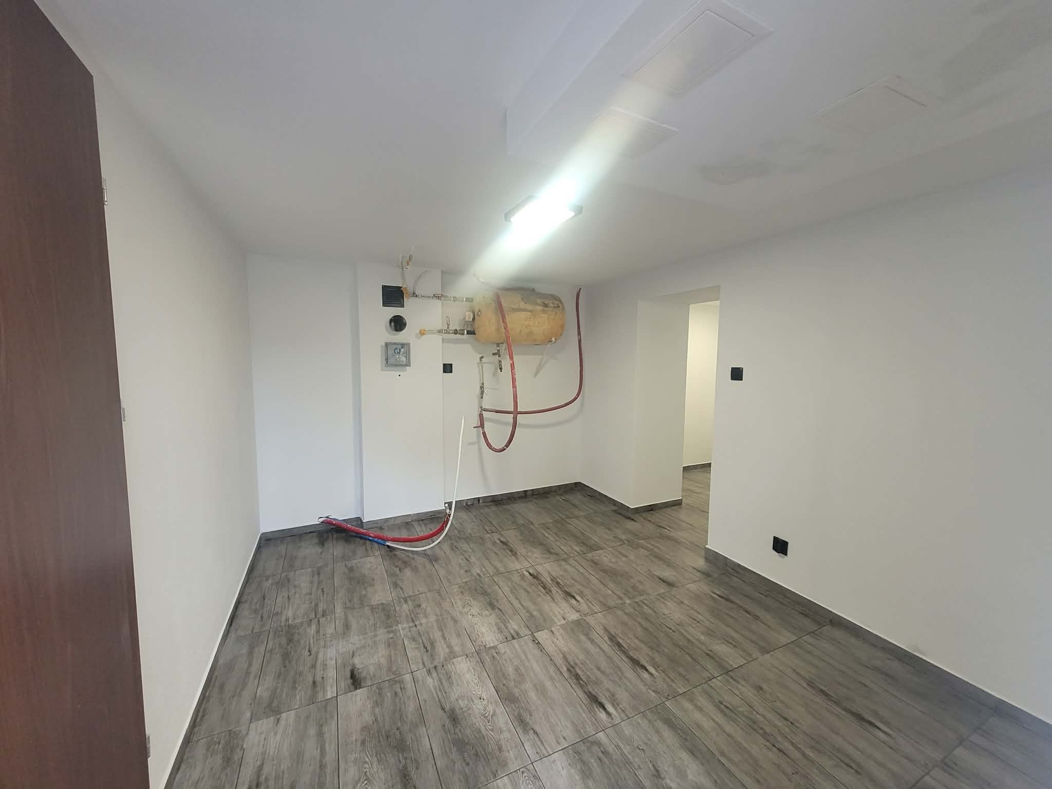 Puste, odświeżone wnętrze z szarymi panelami podłogowymi imitującymi drewno, białymi ścianami i widoczną instalacją grzewczą. Widok na sąsiednie pomieszczenie.