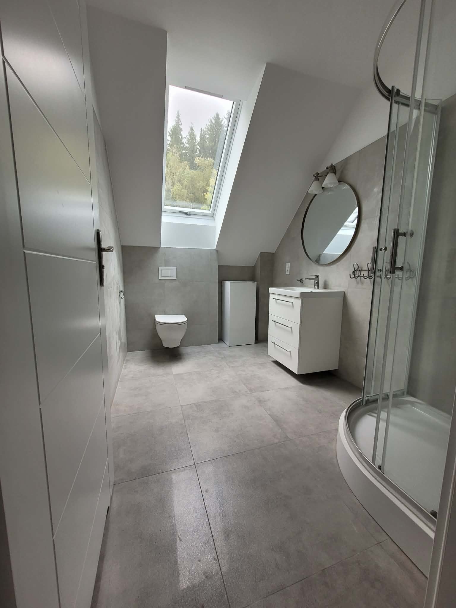 Nowoczesna łazienka na poddaszu z oknem dachowym, szarą glazurą i białą armaturą. Kabina prysznicowa z zaokrąglonymi drzwiami, umywalka z szafką i lustro.