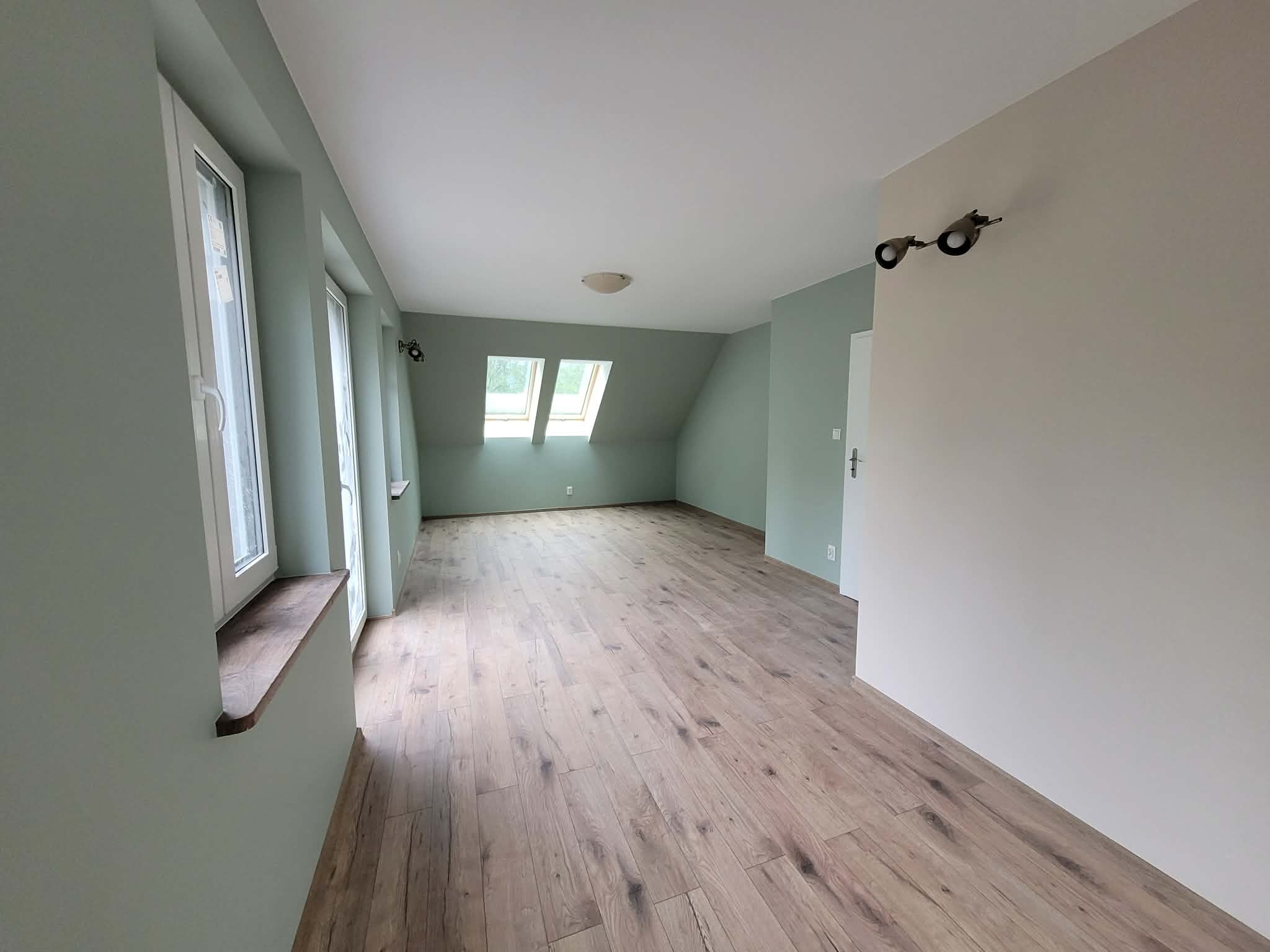 Jasne, przestronne poddasze po remoncie: świeżo pomalowane ściany w odcieniach zieleni, drewnopodobne panele podłogowe, okna dachowe w skosach. Minimalistyczne oświetlenie.