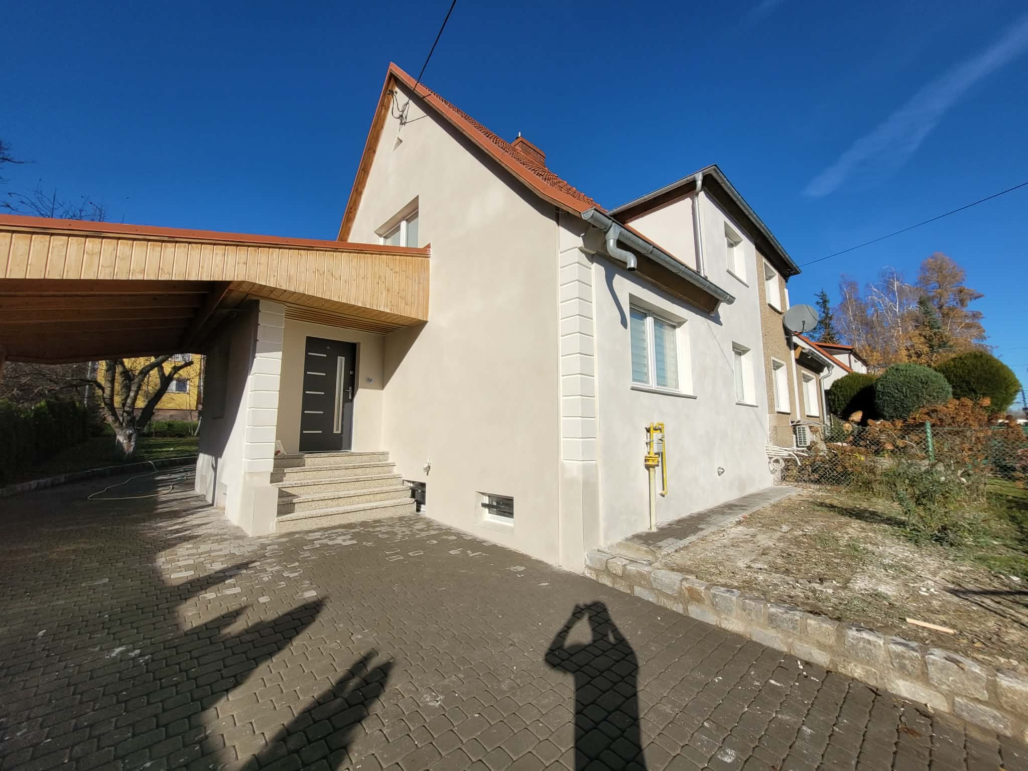 Odnowiona elewacja domu jednorodzinnego z jasnym tynkiem, widoczne detale architektoniczne i podjazd z kostki brukowej. Słoneczny dzień, błękitne niebo.