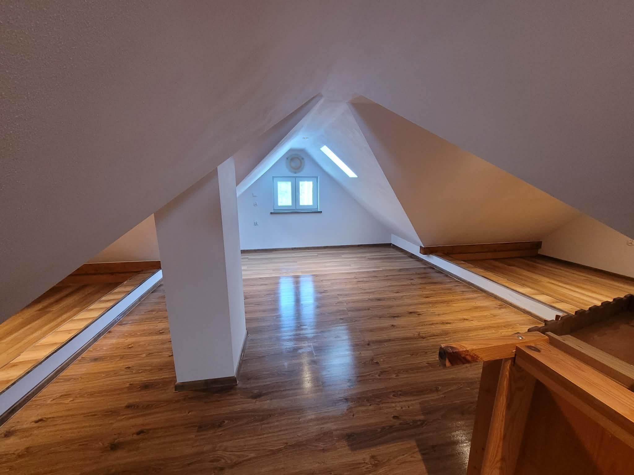 Wykończone poddasze z drewnianą podłogą i białymi ścianami. Widoczne okno dachowe i konstrukcja dachu. Podłoga z wyraźnym wzorem drewna, odbijająca światło.