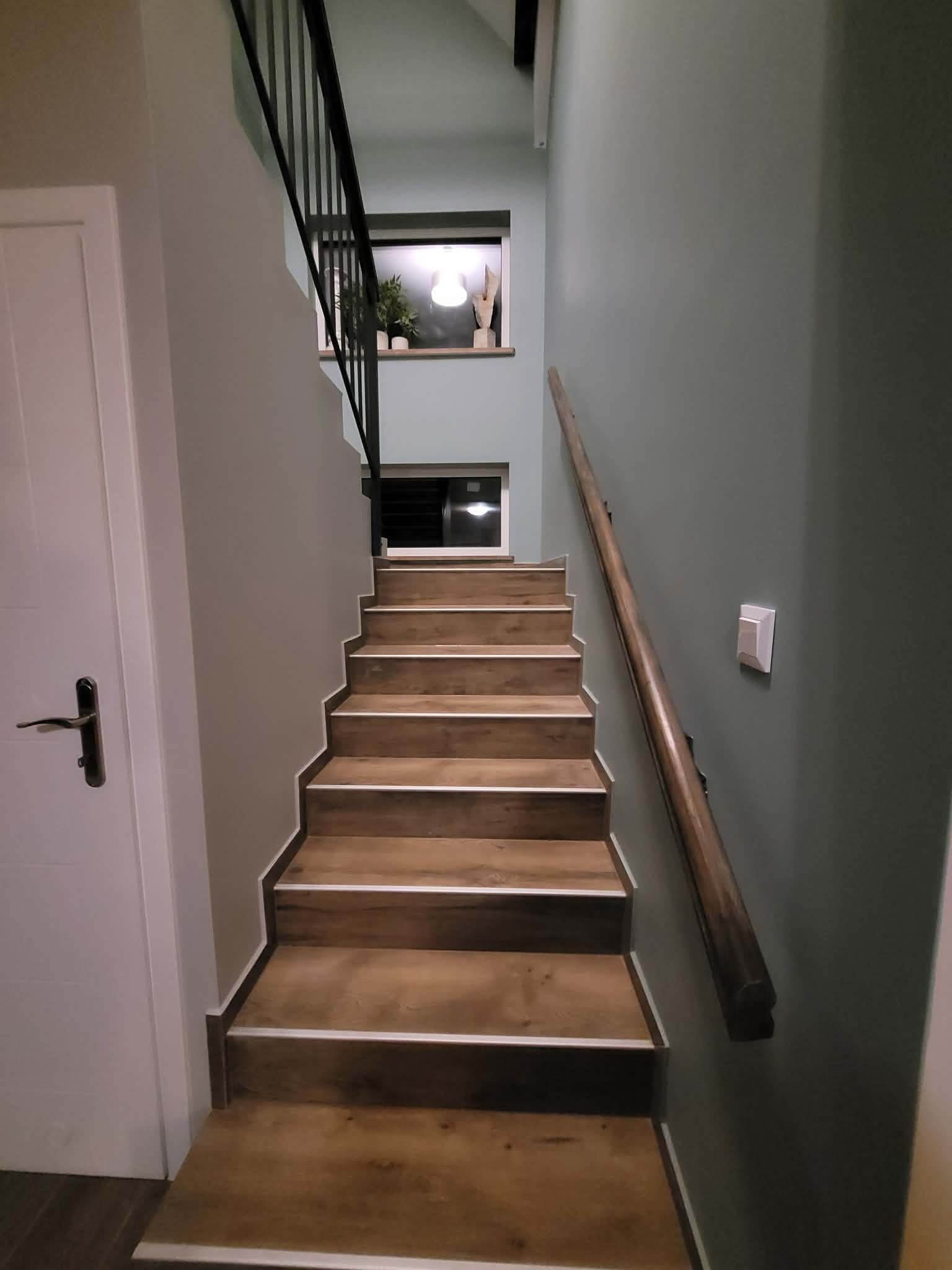 Schody wykończone panelami drewnopodobnymi z białymi listwami, czarna metalowa balustrada, drewniana poręcz na ścianie w odcieniach szarości i zieleni. Widok z dołu.