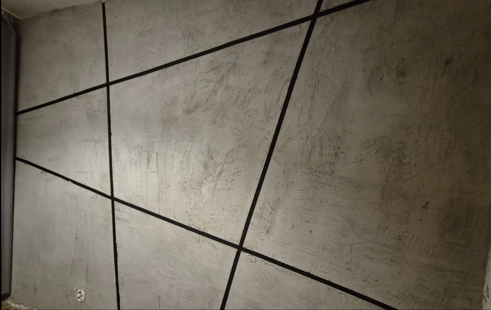 Betonowa ściana z geometrycznym wzorem z czarnych linii, surowy styl, widoczne ślady szpachlowania, gniazdko elektryczne w dolnym rogu.