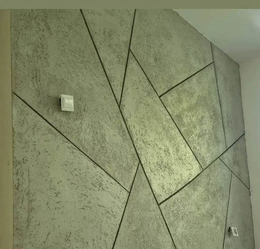 Dekoracyjna ściana z efektem betonu i geometrycznym podziałem na panele. Widoczny włącznik światła i gniazdko elektryczne. Szara kolorystyka.