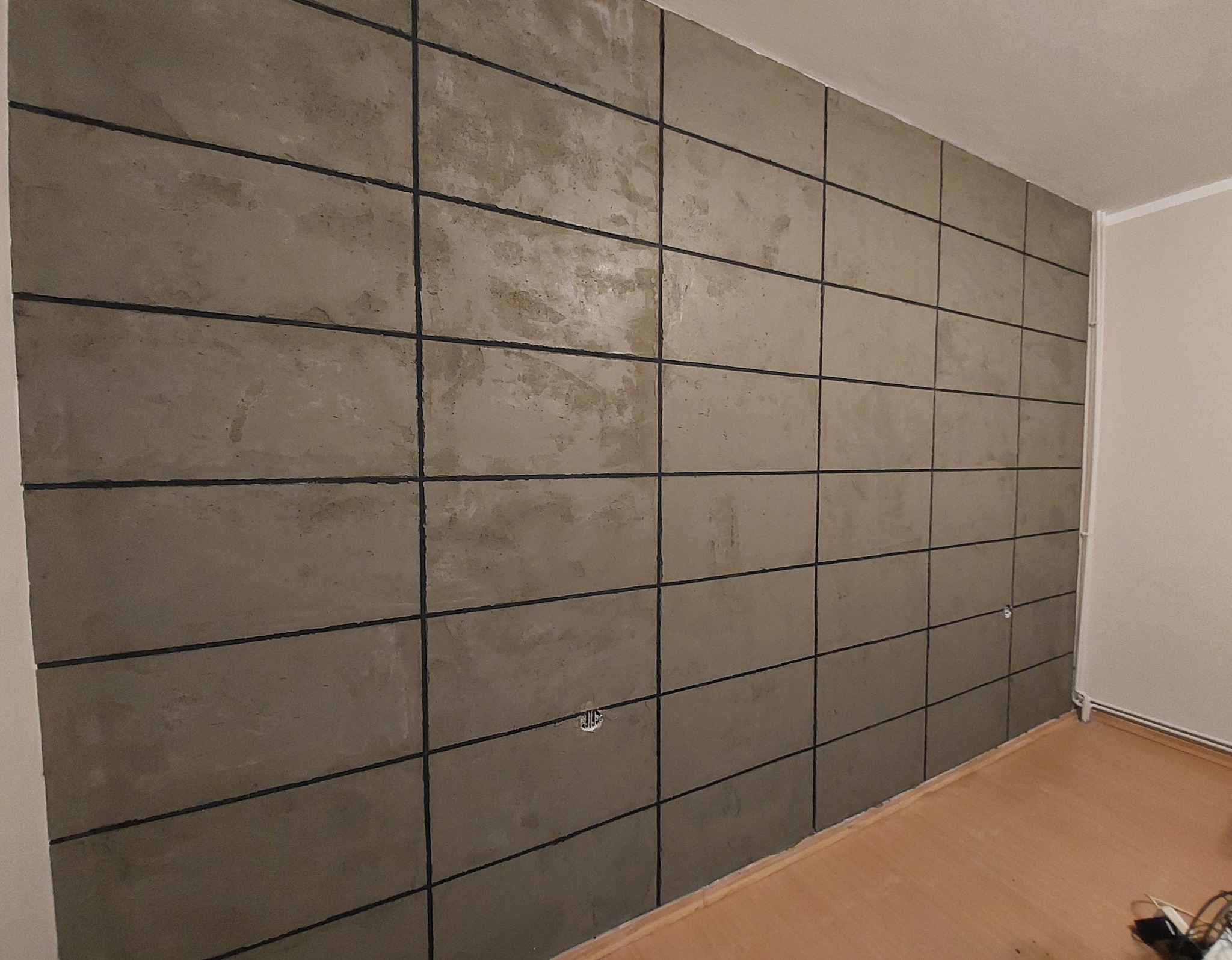 Ściana wykończona w szarych, kwadratowych panelach z ciemnymi fugami, tworzących geometryczny wzór. Widoczne gniazdko elektryczne i fragment podłogi.