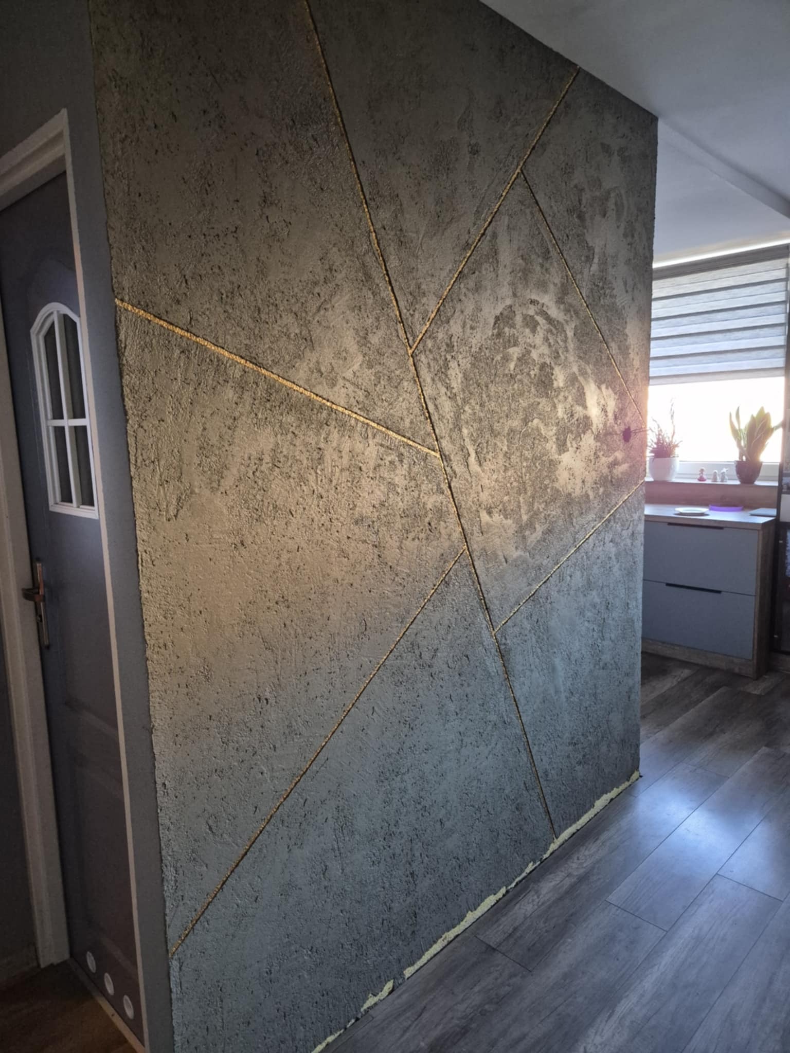 Dekoracyjna ściana w odcieniach szarości z geometrycznym wzorem podkreślonym złotymi liniami, widoczna obok szarych drzwi i fragmentu podłogi.