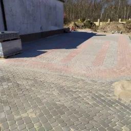 Ułożona nawierzchnia z kostki brukowej w odcieniach szarości i czerwieni, z widocznymi zapasami kostki na paletach i piaskiem między kostkami, na tle budynku i drzew