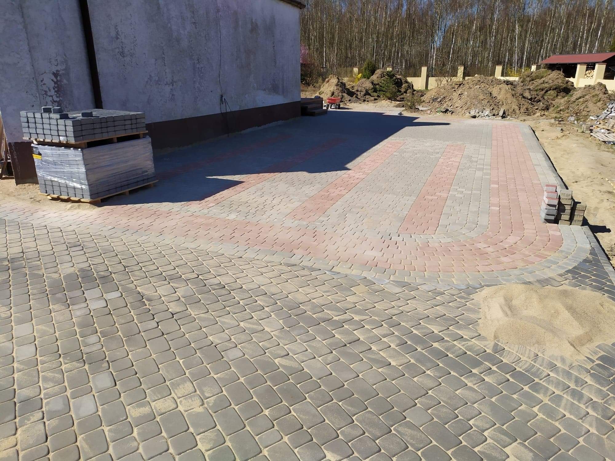 Ułożona nawierzchnia z kostki brukowej w odcieniach szarości i czerwieni, z widocznymi zapasami kostki na paletach i piaskiem między kostkami, na tle budynku i drzew