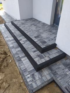 Schody wejściowe wyłożone szarą kostką brukową o nieregularnym wzorze, z czarnymi obrzeżami, prowadzące do drzwi wejściowych budynku.