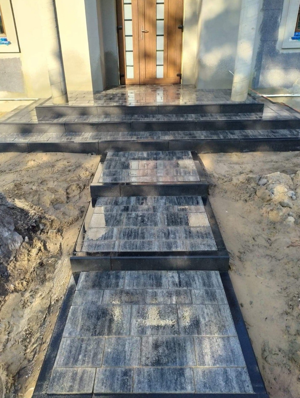 Ułożona kostka brukowa prowadząca do drzwi wejściowych domu, z widocznymi schodami i piaskiem po bokach.