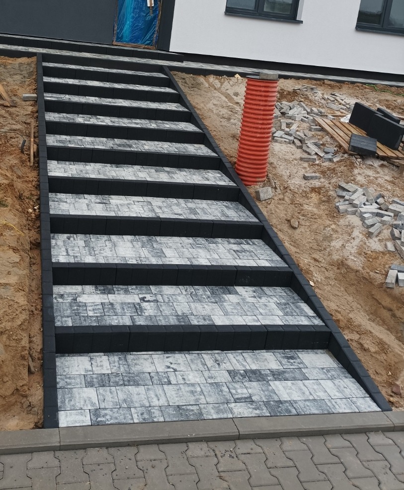 Schody zewnętrzne wyłożone jasnoszarą kostką brukową z czarnymi obrzeżami, prowadzące do budynku z szarą elewacją i oknami, widoczna pomarańczowa rura kanalizacyjna i paleta z kostką w tle.