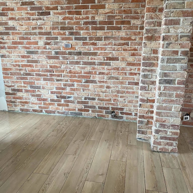 Ściana z czerwonej cegły z wystającą kolumną z cegły, drewniana podłoga z widocznymi słojami, gniazdko elektryczne w ścianie, surowy stan wykończenia.
