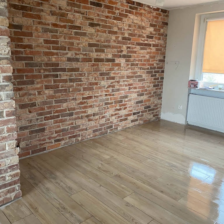 Pokój po remoncie z ceglaną ścianą i jasną drewnopodobną podłogą, widoczny fragment okna z grzejnikiem.