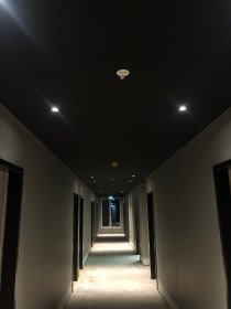 Korytarz z ciemnym sufitem i punktowym oświetleniem LED, widok w perspektywie, drzwi po obu stronach, światło dzienne wpadające z końca korytarza.