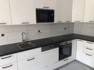 Nowoczesna, biała kuchnia z czarnym blatem i srebrnymi sprzętami AGD. Widoczne płytki ceramiczne na ścianie, jeszcze nie w pełni wykończone otwory na gniazdka elektryczne. Minimalistyczne uchwyty...