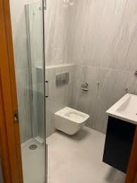 Nowoczesna łazienka z kabiną prysznicową z przezroczystego szkła, podwieszaną miską WC z chromowanym przyciskiem spłukiwania, umywalką nablatową na czarnej szafce i jasnymi płytkami imitującymi...