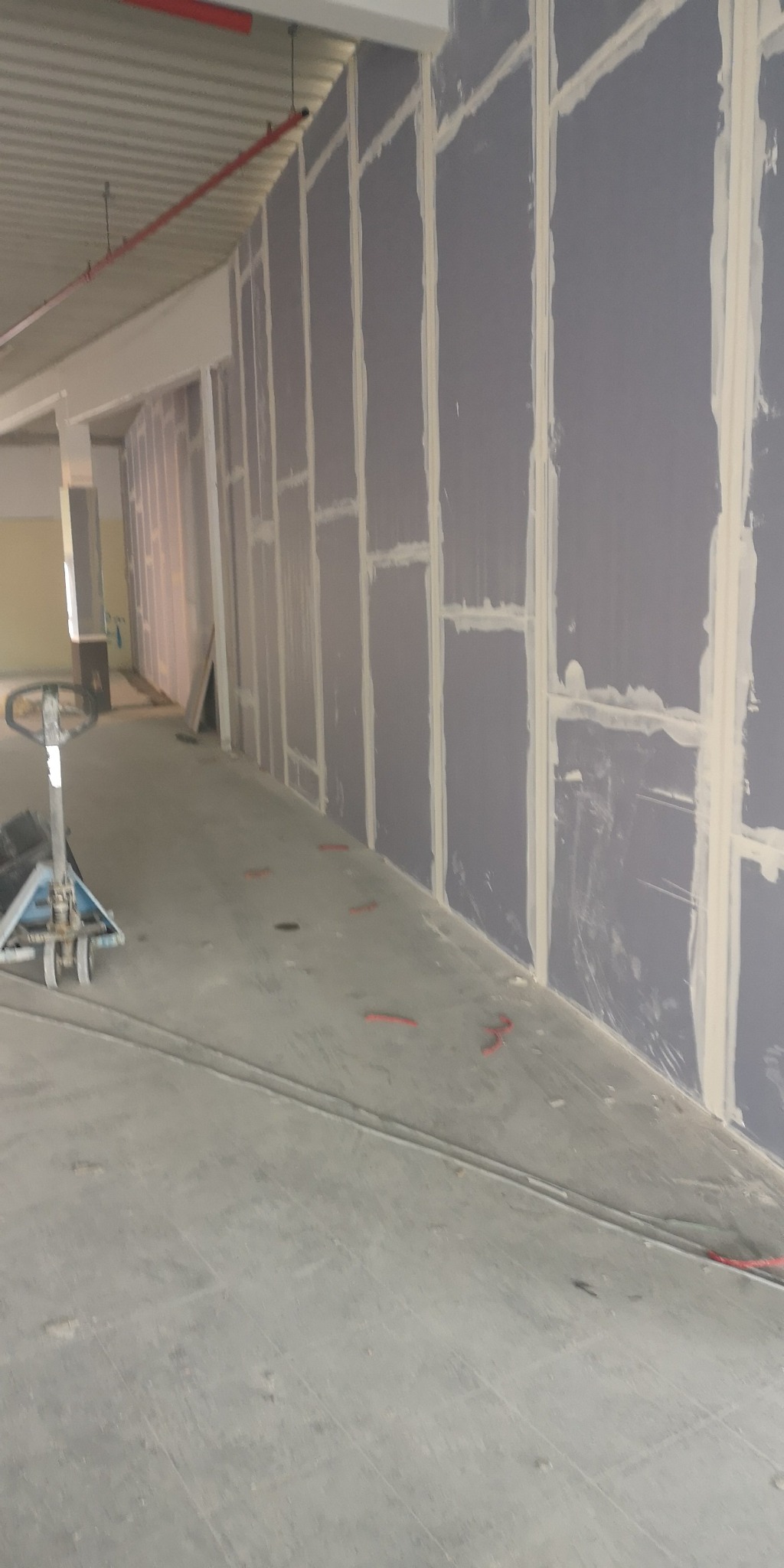 Montaż płyt karton gips na ścianie w hali, widoczne łączenia płyt wypełnione masą szpachlową, podłoga betonowa z czerwonymi oznaczeniami.