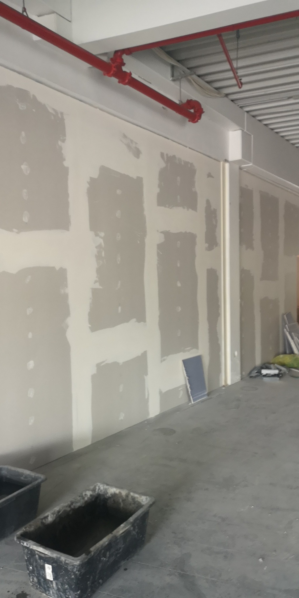 Przygotowana ściana z płyt kartonowo-gipsowych, z widocznymi łączeniami zaszpachlowanymi na gładko, czerwona instalacja rurowa pod sufitem, plastikowe pojemniki na podłodze.
