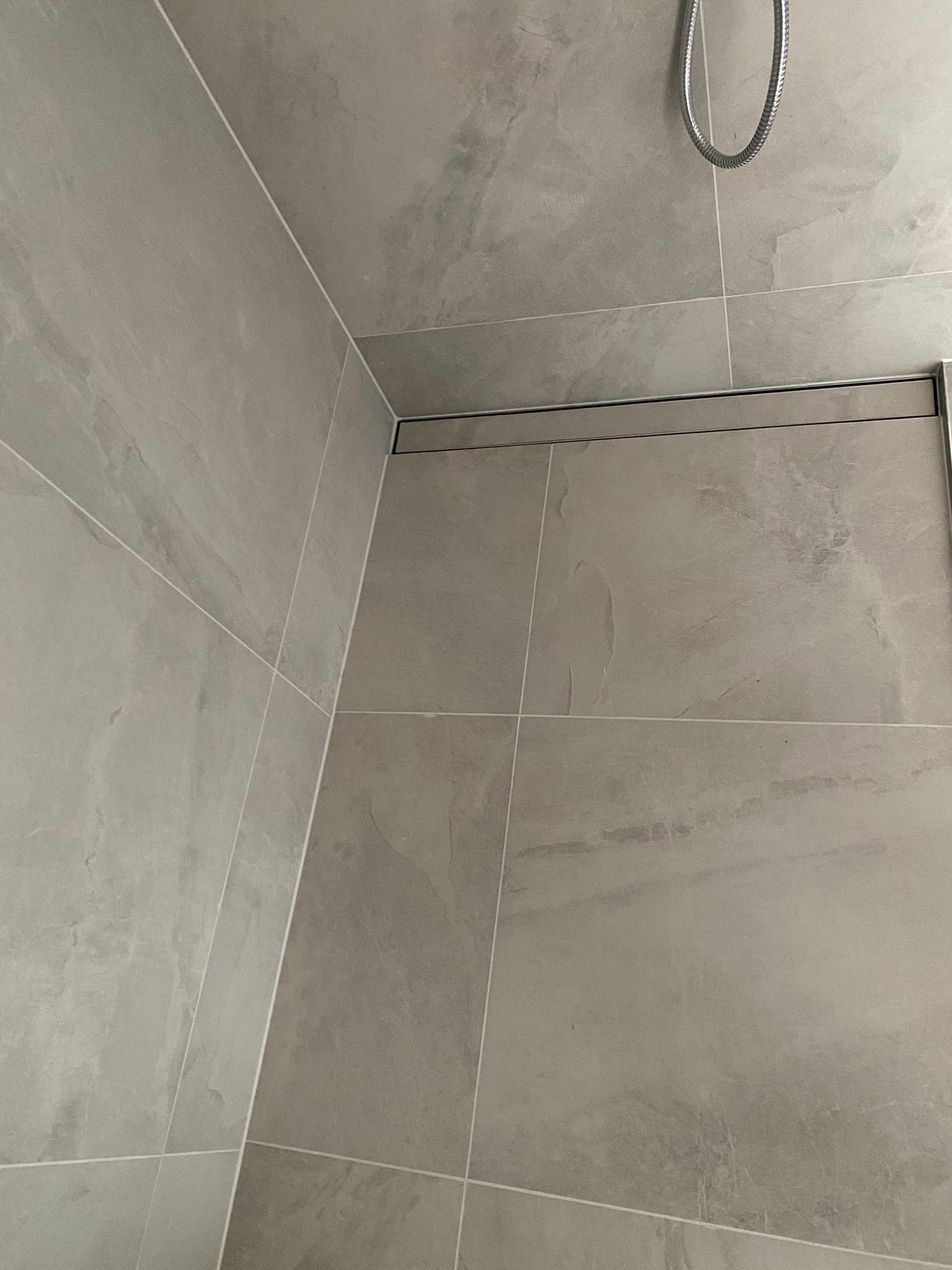 Szare płytki imitujące kamień w kabinie prysznicowej z odpływem liniowym i słuchawką prysznicową.