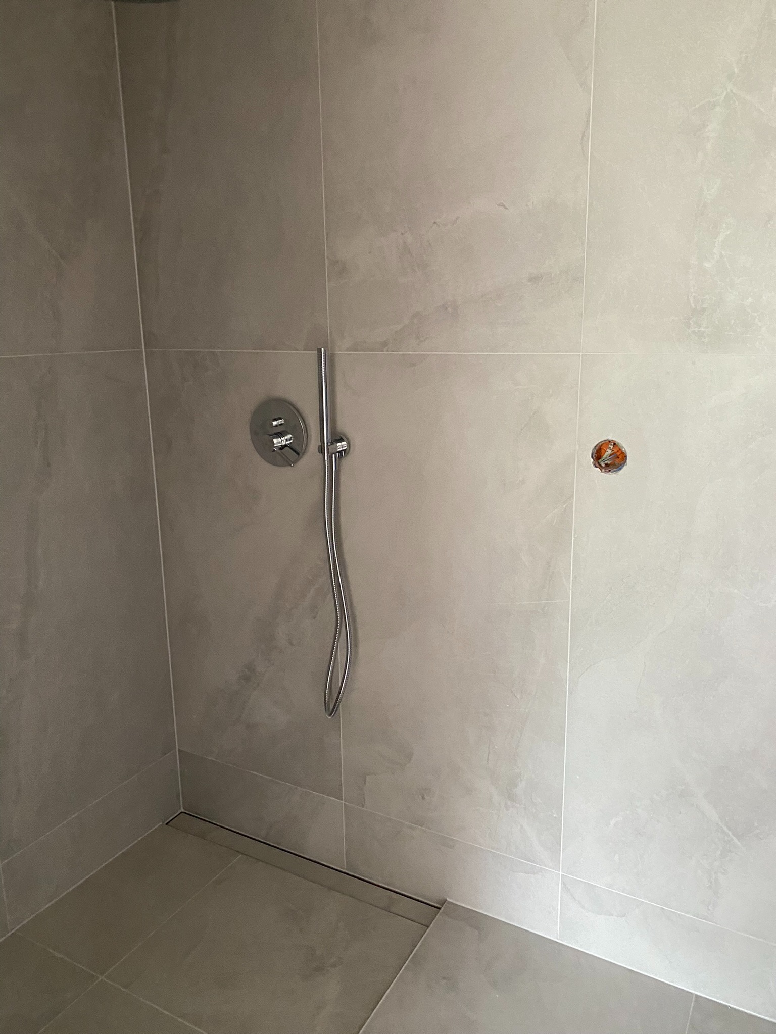 Szara łazienka w trakcie wykańczania, widoczny panel prysznicowy z termostatem, odpływ liniowy w podłodze i niedokończone podłączenie elektryczne.