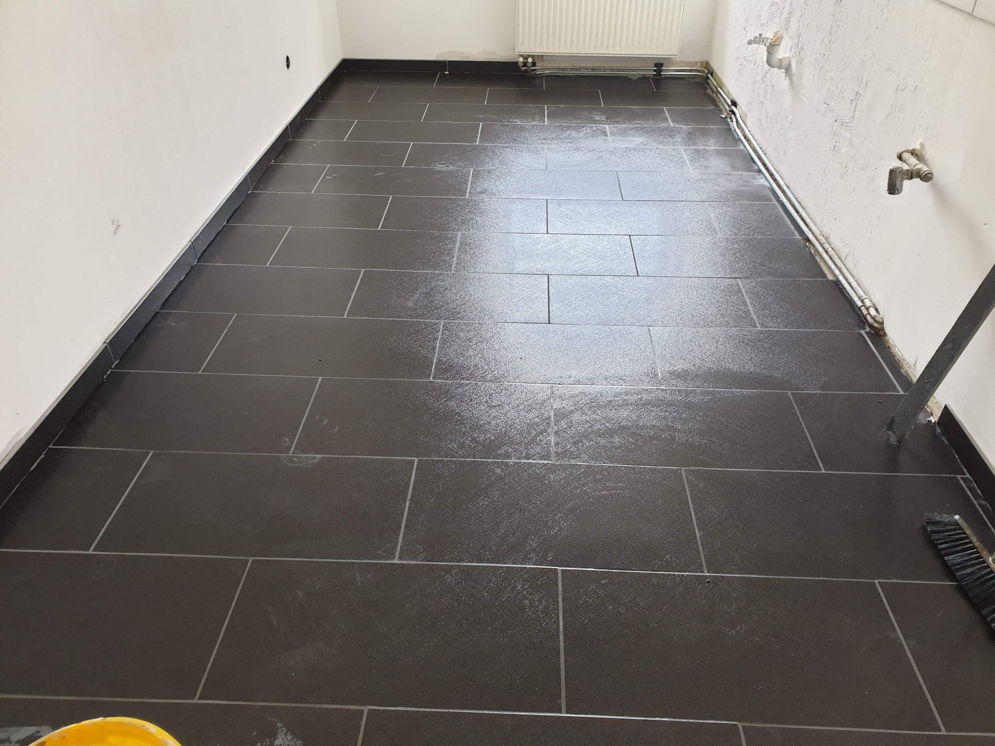Ułożona, czarna, prostokątna podłoga z płytek, świeżo po umyciu, z widocznymi rurami grzewczymi przy ścianie i fragmentem szczotki do sprzątania.