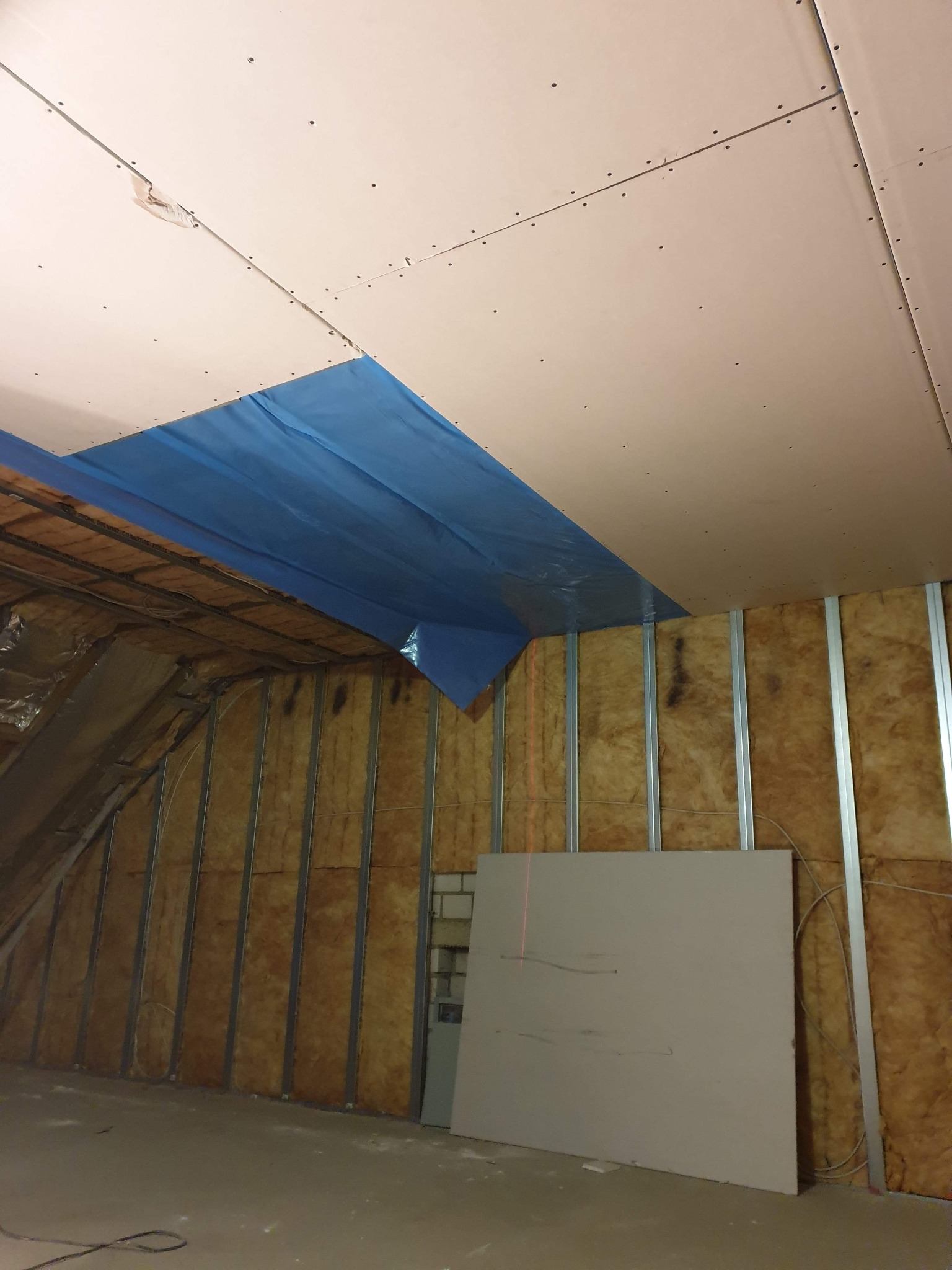 Poddasze w trakcie prac wykończeniowych: izolacja ścian wełną mineralną, montaż metalowych profili, częściowo zamontowane płyty gipsowo-kartonowe na suficie z zabezpieczeniem folią, widoczny laser...