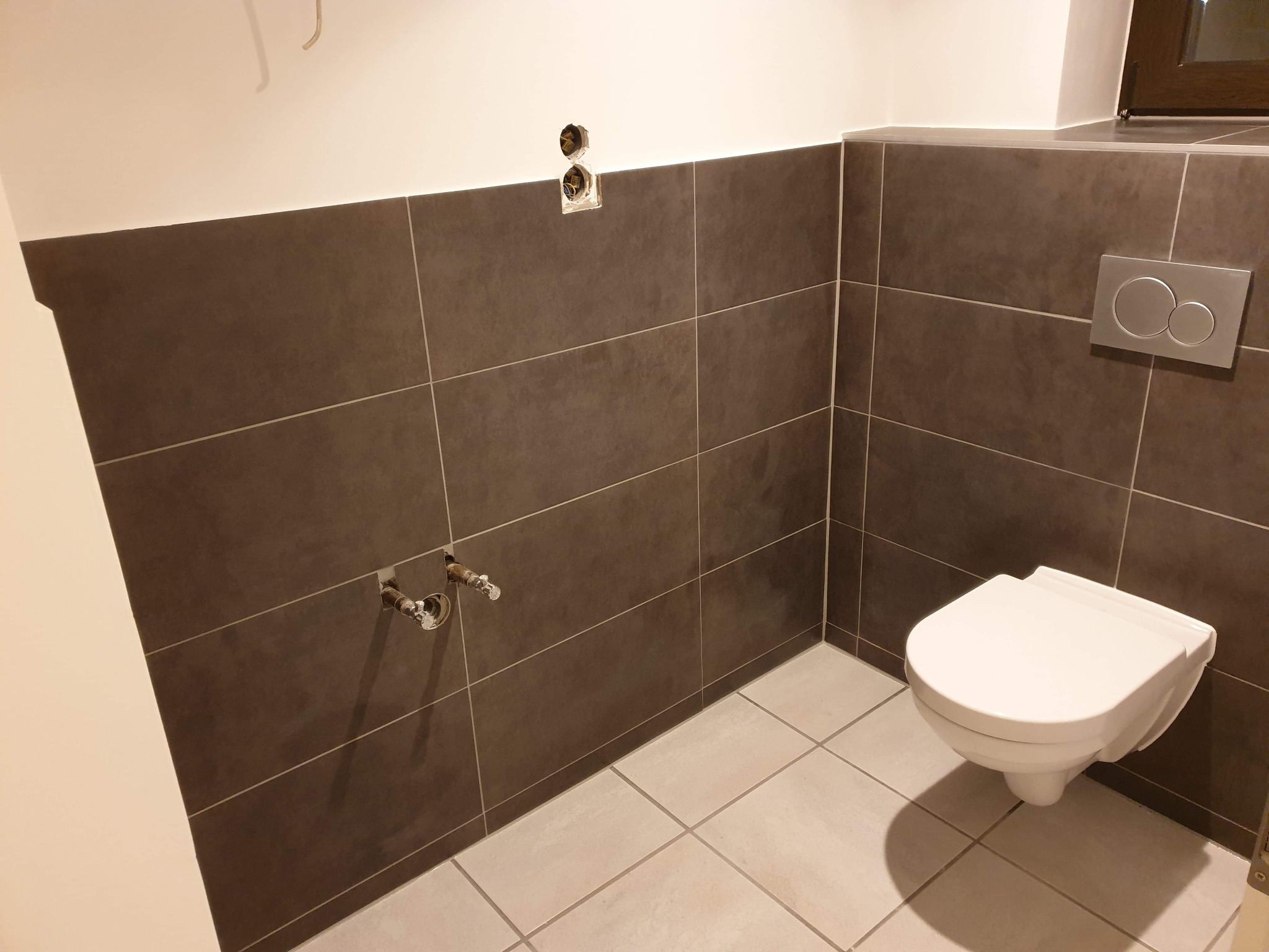 Świeżo wykafelkowana łazienka z wiszącą toaletą, widoczne niezabezpieczone wyprowadzenia rur i gniazdka elektryczne przed montażem osprzętu.