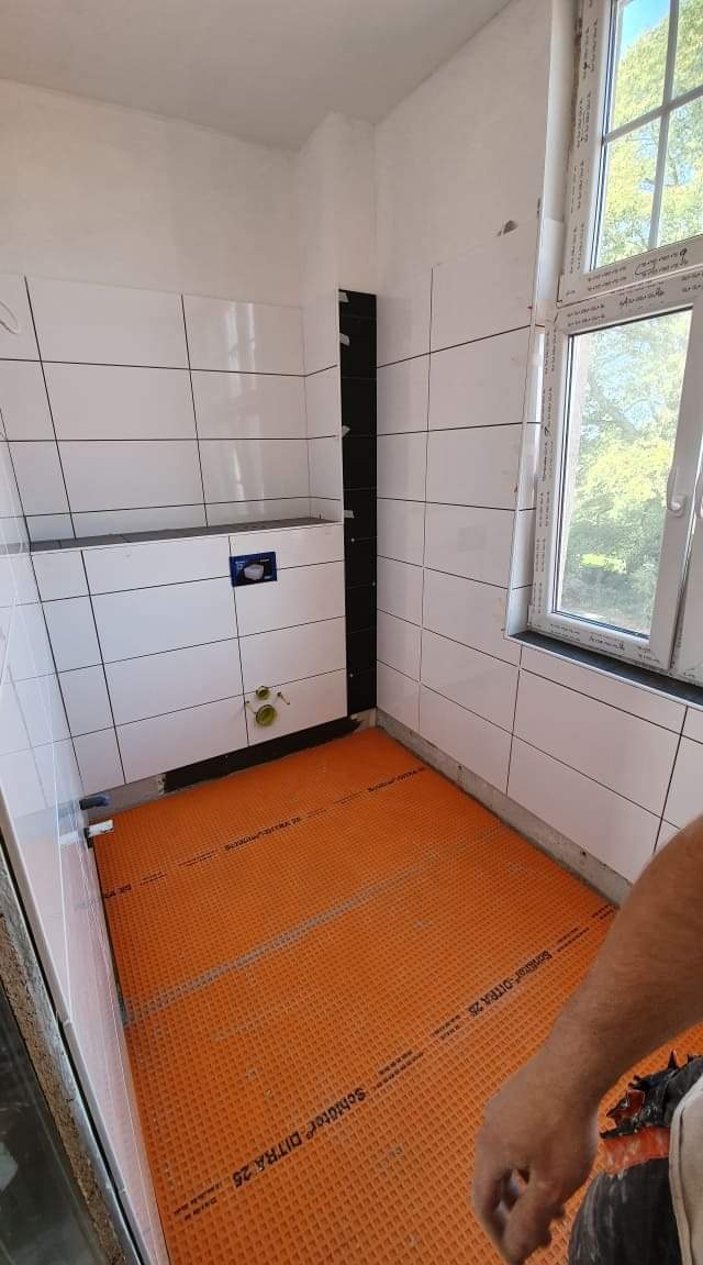 Remont łazienki w trakcie: białe płytki ścienne, czarny pas płytek, pomarańczowa mata izolacyjna Schluter DITRA 25 na podłodze, okno z widokiem na zieleń na zewnątrz.