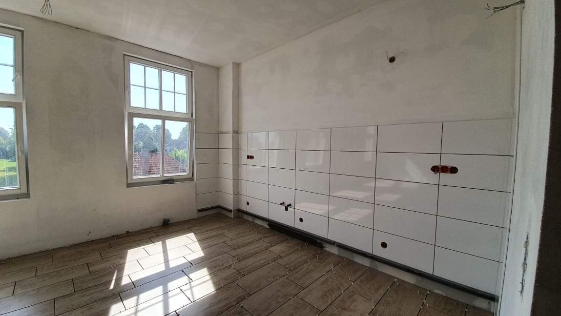 Wnętrze w trakcie układania płytek podłogowych i ściennych; widoczne białe płytki na ścianach z przygotowanymi otworami na gniazdka i wyprowadzenia rur, oraz ułożone płytki podłogowe w odcieniach...