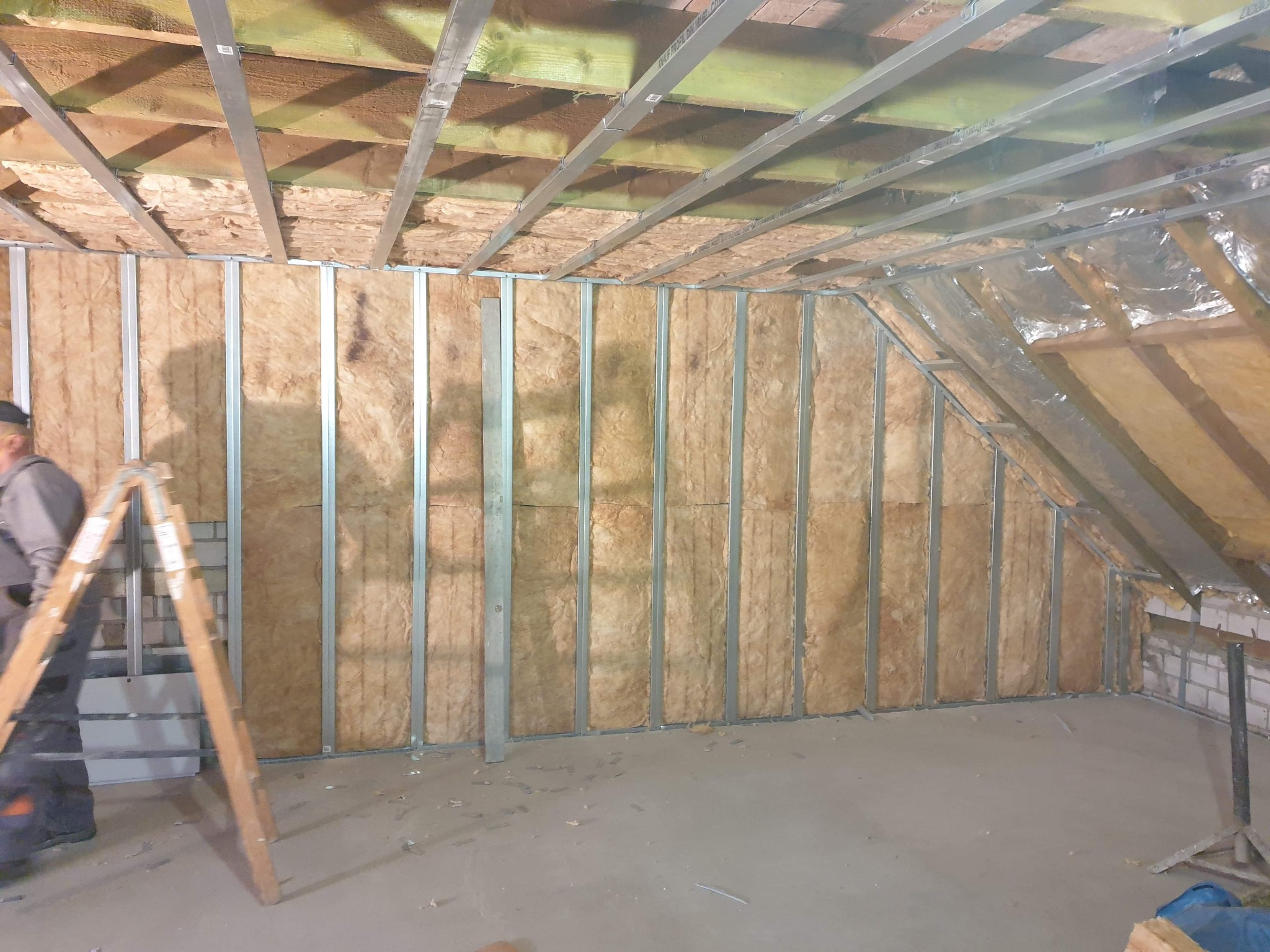Poddasze w trakcie izolacji: wełna mineralna między metalowymi profilami konstrukcyjnymi, widoczna drabina i częściowo widoczny pracownik.