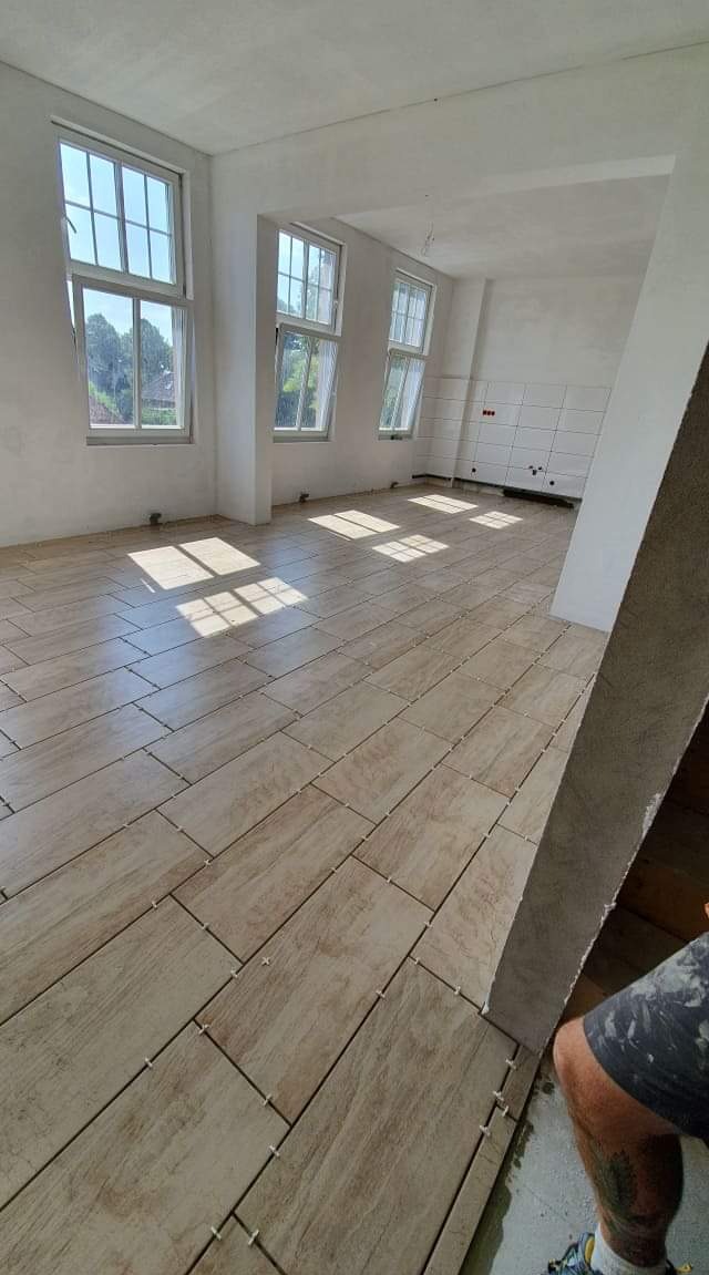 Układanie podłogi z płytek imitujących drewno w jasnym, słonecznym pokoju z trzema oknami; widoczne krzyżyki dystansowe i fragment nogi glazurnika.