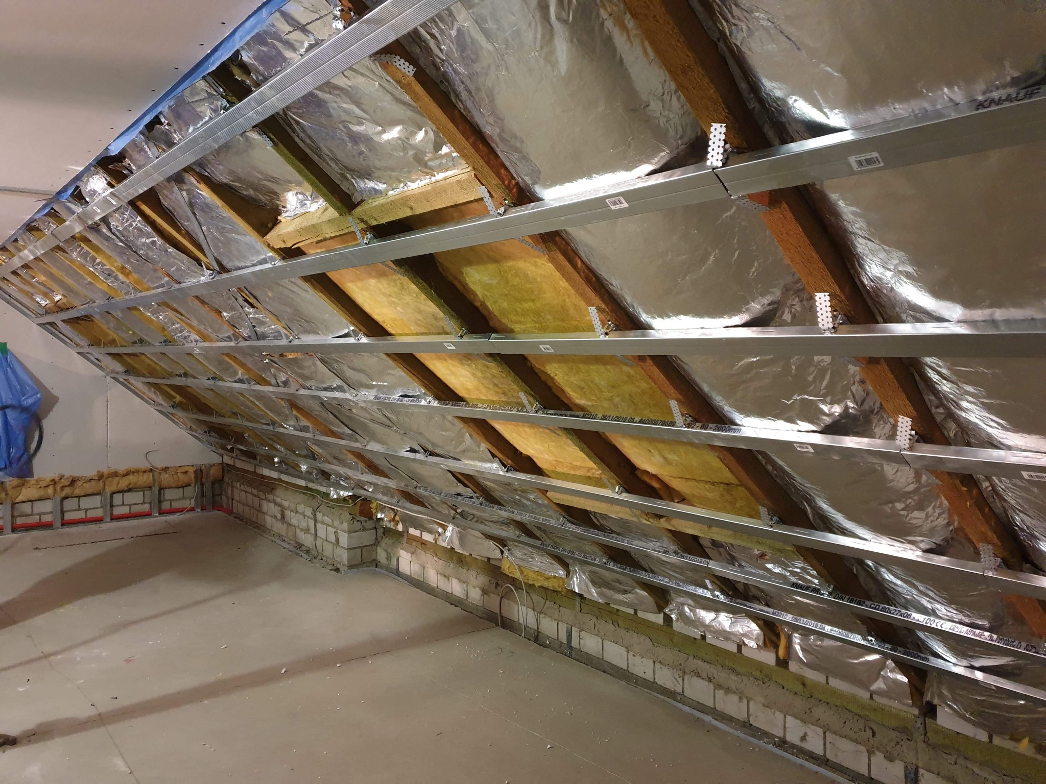 Poddasze w trakcie adaptacji: drewniana konstrukcja dachu ocieplona wełną mineralną i folią aluminiową, zamontowane metalowe profile pod płyty kartonowo-gipsowe, widoczne fragmenty muru i instalacji.